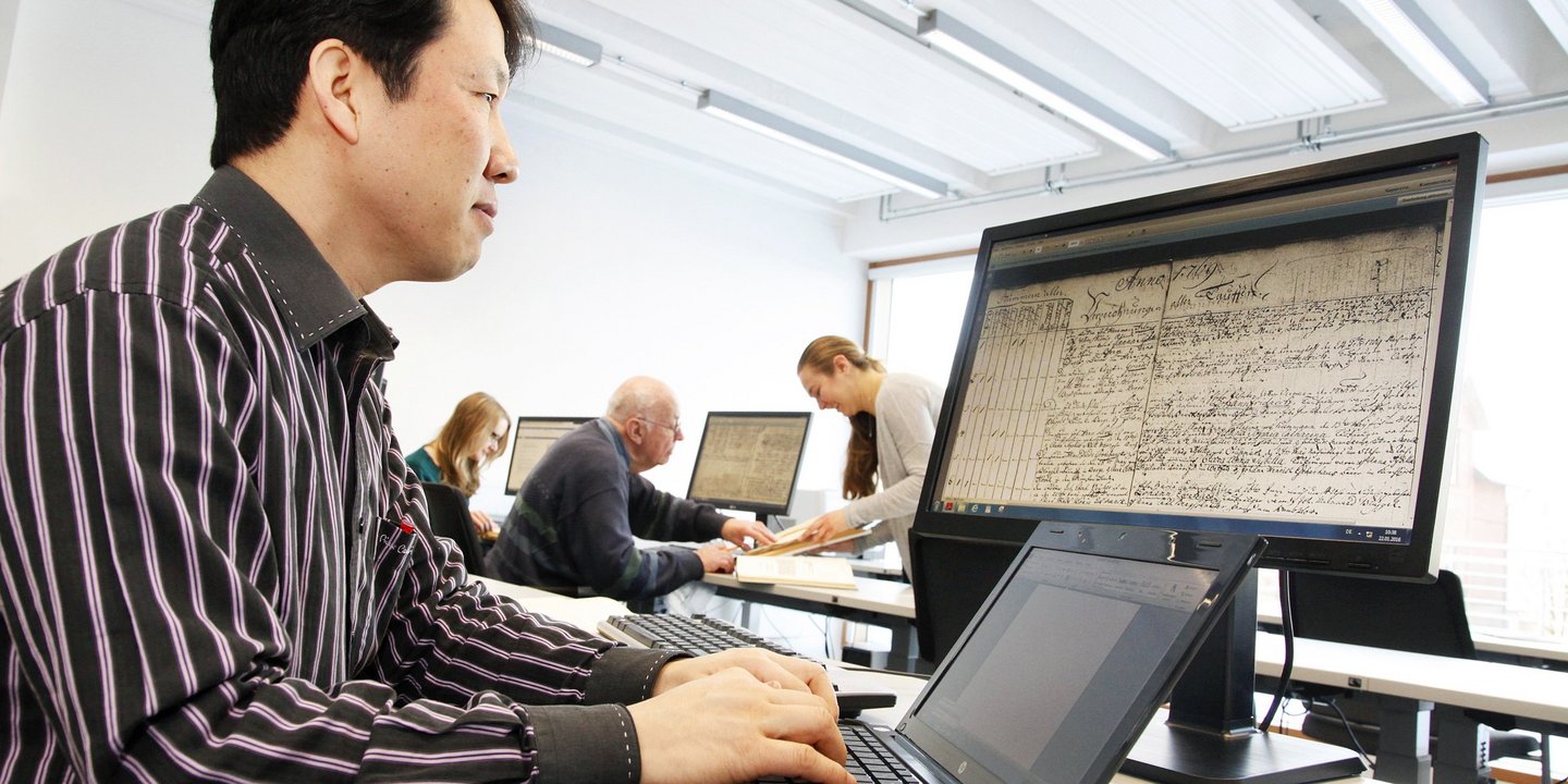 Ein Mann betrachtet ein altes Dokument am PC Bildschirm und arbeitet am Laptop